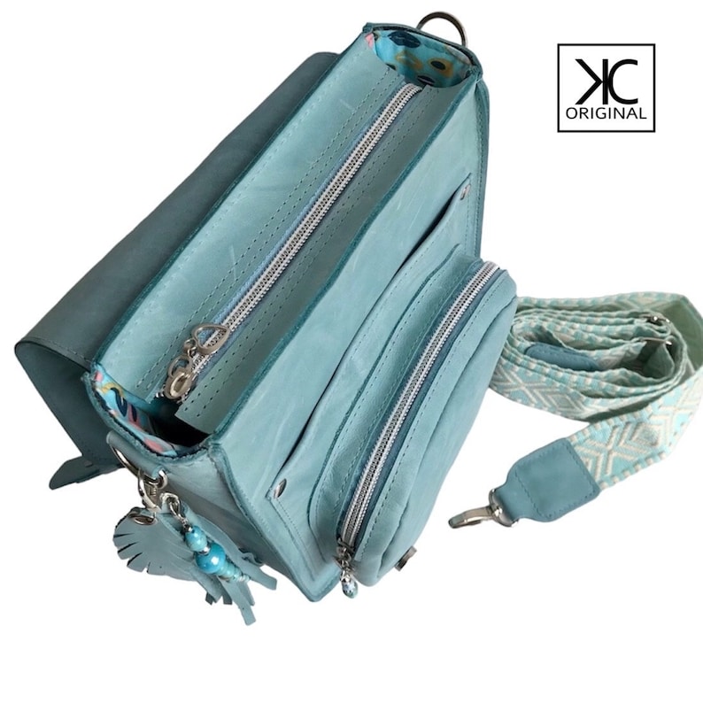 May include: A light blue leather crossbody bag with a zippered closure and a patterned strap. The bag has a small charm attached to it. The bag is open and the inside is lined with a floral pattern. The bag is labeled "KC ORIGINAL".