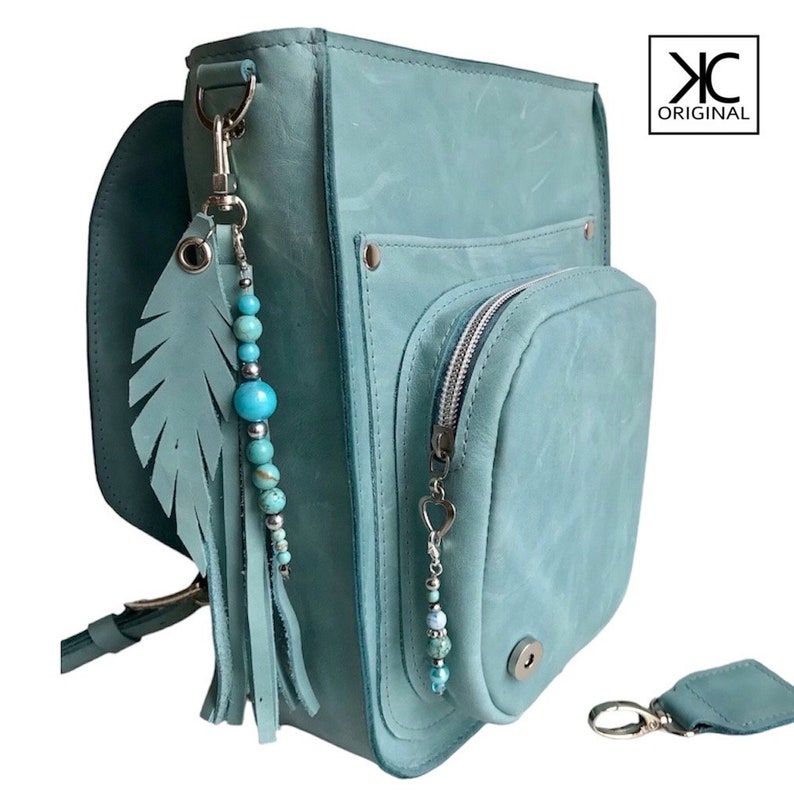 May include: A teal blue leather crossbody bag with a zippered pocket and a silver zipper pull. The bag has a leather strap and a turquoise feather charm with beads.