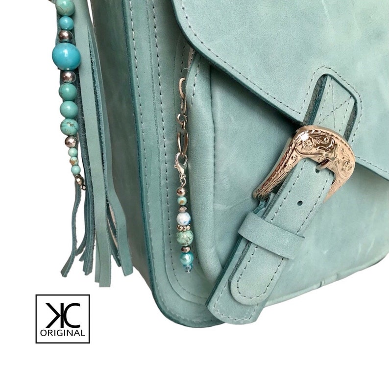 May include: A light blue leather crossbody bag with a silver buckle and a beaded tassel. The bag has a flap closure and a zippered pocket on the front.