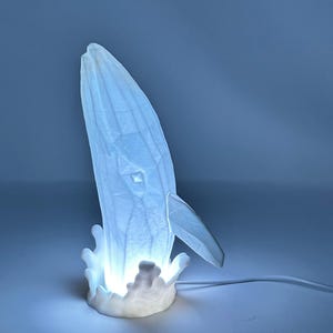 May include: A white 3D printed whale lamp with a light blue glow, leaping out of the water. The whale is detailed with fins and a tail, and the water is depicted with a splash of waves.