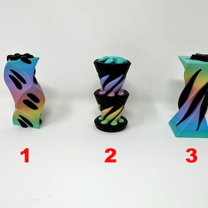 May include: Three 3D printed rainbow colored objects. The first object is a single piece with a wavy design. The second object is a two-piece stack with a spiral design. The third object is a single piece with a spiral design.
