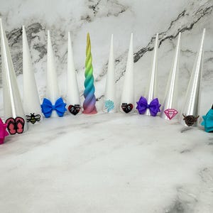 May include: A collection of decorative unicorn horns in various colors and designs. The horns are white, black, and rainbow-colored, with bows and embellishments. The bows are pink, blue, and purple. Some horns have small decorative charms.