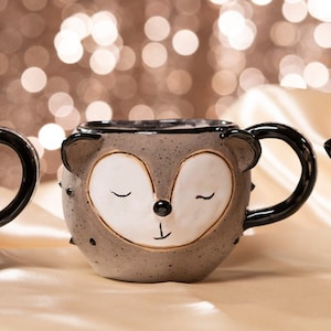 Può includere: Tre tazze in ceramica con facce di animali. Le tazze hanno una base grigia con manici e dettagli neri. Ogni tazza presenta una faccia di animale diversa: una volpe, un riccio e un panda, tutti con gli occhi chiusi e un'espressione sorridente.
