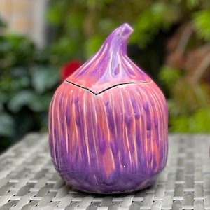 Peut inclure: Un récipient en céramique en forme de figue avec un glaçage violet et rose. Le récipient a un couvercle et est posé sur une surface tissée.