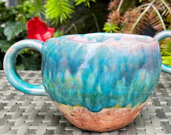 Könnte beinhalten: Zwei handgefertigte Keramikbecher mit einer einzigartigen türkisfarbenen, blauen und rosa Glasur. Die Becher haben ein rustikales, strukturiertes Aussehen mit einem braunen Boden und gebogenen Griffen. Perfekt für Kaffee oder Tee.