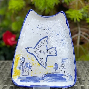 Könnte beinhalten: Eine weiße Keramik-Servierplatte in Katzenform mit blauem Rand. Die Platte zeigt ein handgemaltes Design einer Taube mit einem Olivenzweig, die über einer blauen und gelben Landschaft mit Bäumen und einem kleinen Haus fliegt.