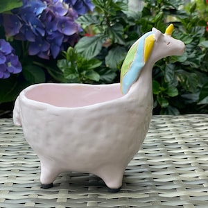 May include: A pink ceramic planter shaped like a unicorn with a rainbow mane. The planter has a small opening at the top for planting.
