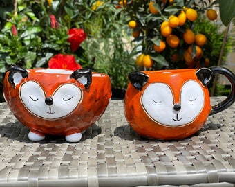 Könnte beinhalten: Zwei Keramikbecher in Form eines Fuchses, einer mit Henkel und einer ohne. Die Becher sind orange mit schwarzen Ohren und weißen Gesichtern. Sie stehen auf einer gewebten Oberfläche.