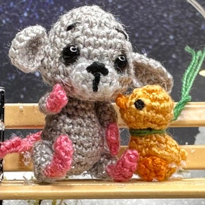 May include: Crocheted gray mouse and orange puppy dolls sitting on a miniature wooden bench. The mouse has pink feet and a long pink tail. The puppy has a green collar. The background is a blurred image of a starry night.