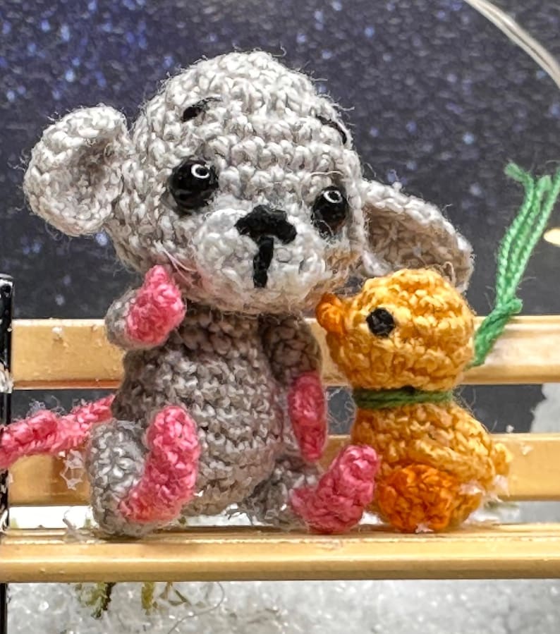 May include: Crocheted gray mouse and orange puppy dolls sitting on a miniature wooden bench. The mouse has pink feet and a long pink tail. The puppy has a green collar. The background is a blurred image of a starry night.