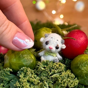 May include: A miniature white and gray cow figurine with a green scarf, nestled among green kale, a red radish, and brussels sprouts. The cow has black eyes and pink accents. A hand with a pink and white manicure is in the frame.