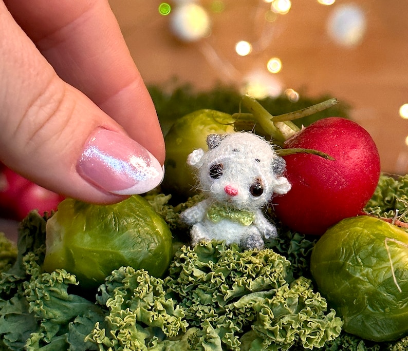 May include: A miniature white and gray cow figurine with a green scarf, nestled among green kale, a red radish, and brussels sprouts. The cow has black eyes and pink accents. A hand with a pink and white manicure is in the frame.