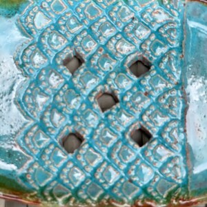 May include: A turquoise ceramic soap dish with a decorative, geometric pattern and cut-out holes. The dish has a glossy finish and a brown rim.