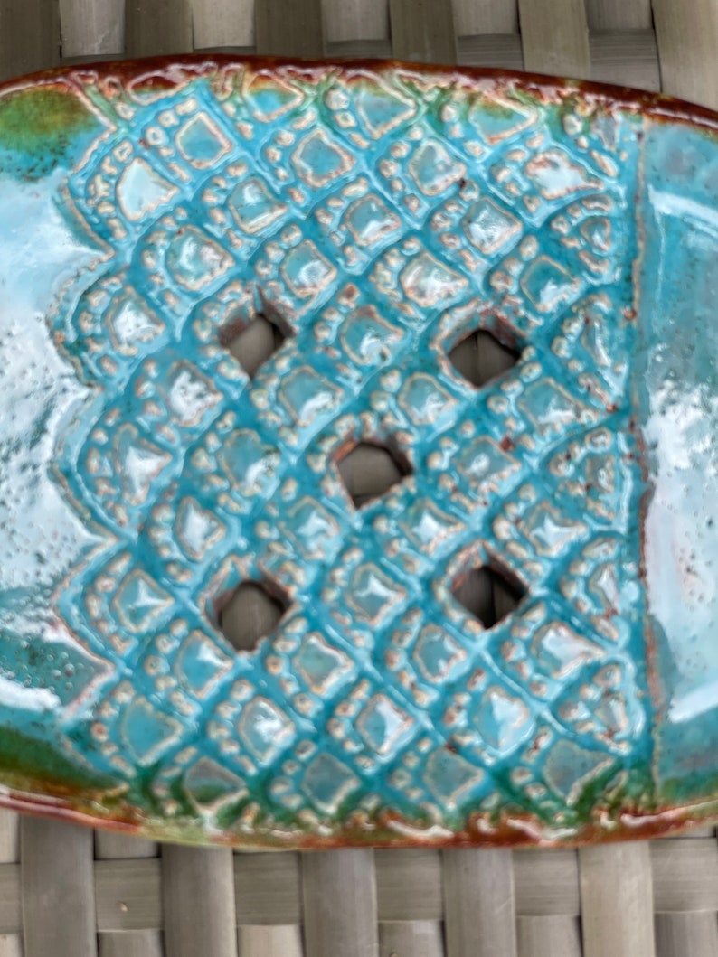 May include: A turquoise ceramic soap dish with a decorative, geometric pattern and cut-out holes. The dish has a glossy finish and a brown rim.