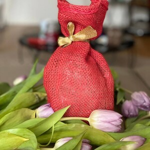 May include: A red burlap gift bag with a gold ribbon tied around the top. The bag is sitting on a bed of purple tulips.