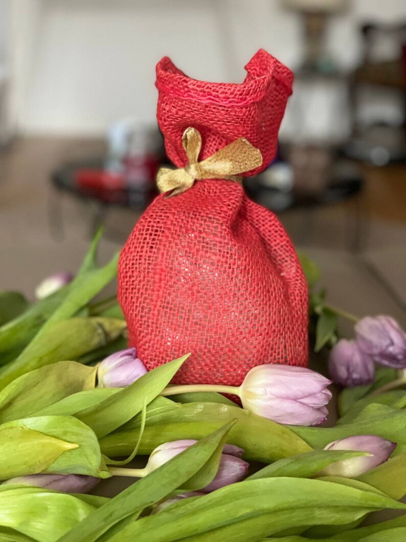 May include: A red burlap gift bag with a gold ribbon tied around the top. The bag is sitting on a bed of purple tulips.