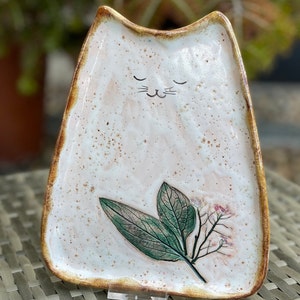 May include: A white ceramic cat-shaped dish with a brown rim and a painted floral design. The dish has a cute cat face with closed eyes and whiskers.