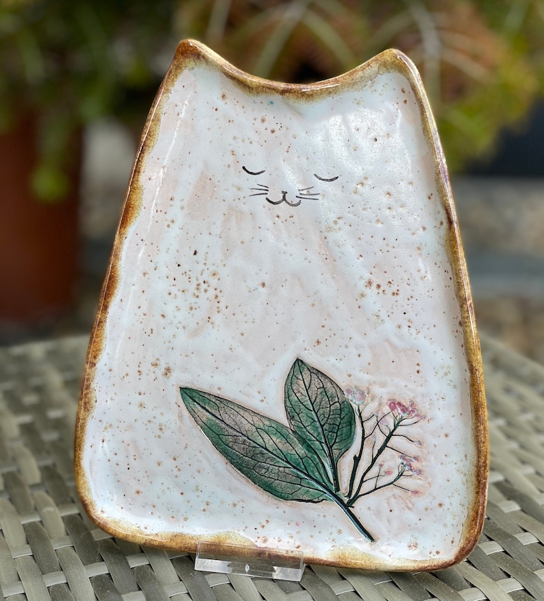 May include: A white ceramic cat-shaped dish with a brown rim and a painted floral design. The dish has a cute cat face with closed eyes and whiskers.