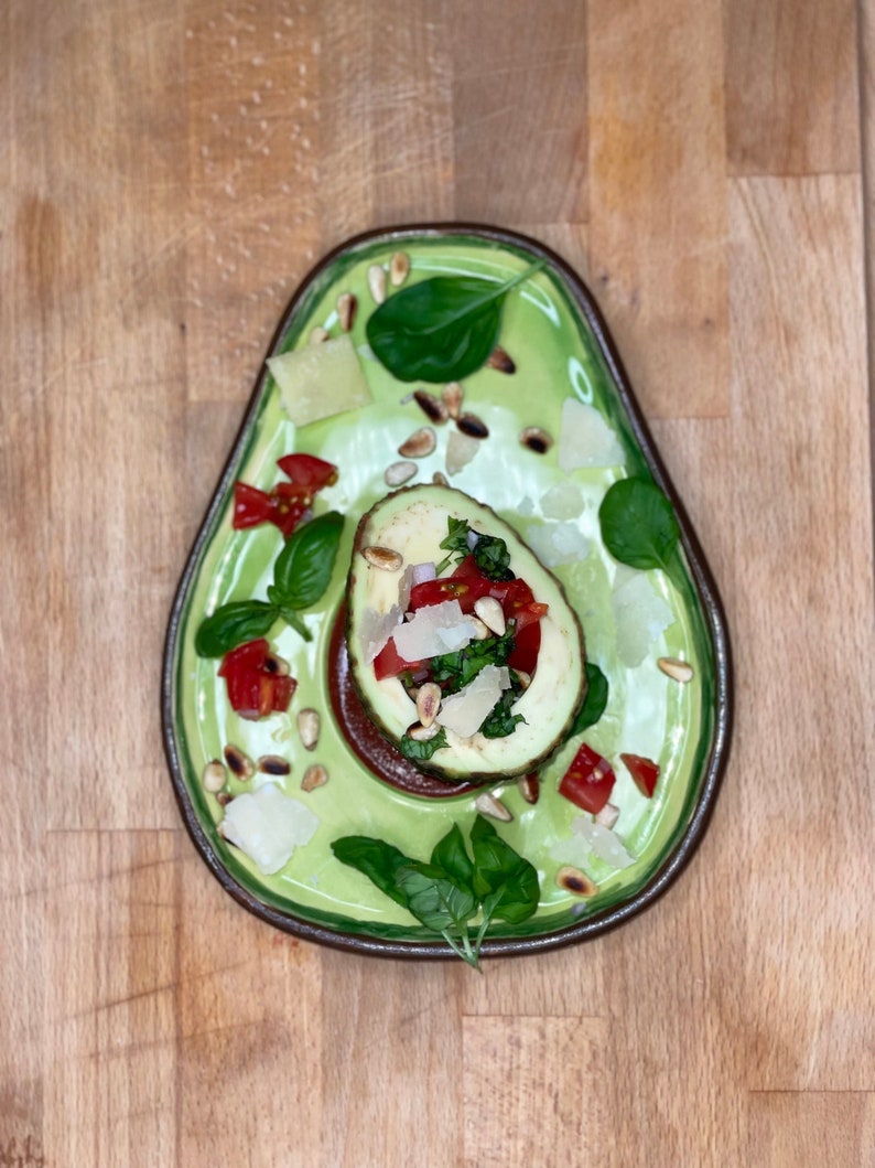 May include: A green ceramic plate shaped like an avocado with a halved avocado filled with chopped tomatoes, pine nuts, basil, and parmesan cheese.