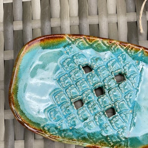 May include: A turquoise ceramic soap dish with a textured design and four drainage holes. The dish has a brown and green glaze around the edges.