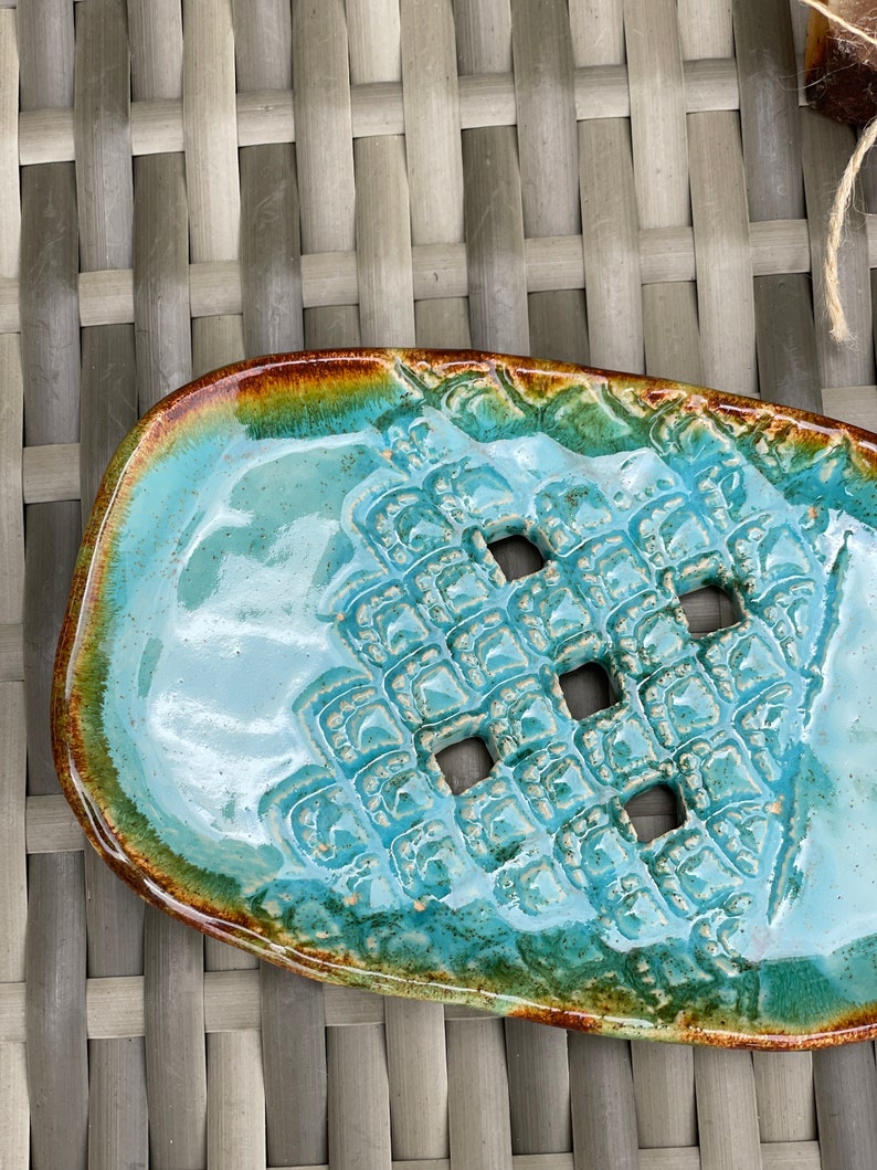 May include: A turquoise ceramic soap dish with a textured design and four drainage holes. The dish has a brown and green glaze around the edges.