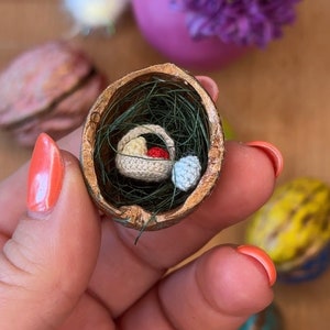 May include: A miniature crochet basket with yellow and red items inside, nestled in a walnut shell with green faux grass. A small blue crochet item sits beside the basket. The shell is held in a hand.