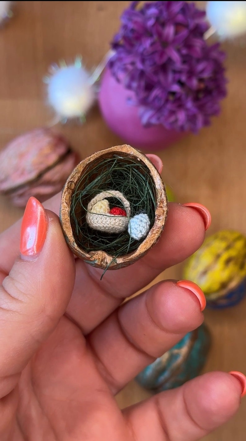 May include: A miniature crochet basket with yellow and red items inside, nestled in a walnut shell with green faux grass. A small blue crochet item sits beside the basket. The shell is held in a hand.