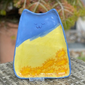 May include: A blue and yellow ceramic dish shaped like a cat with a sleeping face. The dish is decorated with yellow sunflowers around the edge.