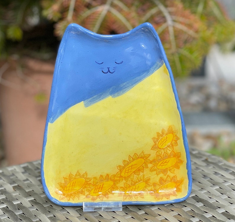 May include: A blue and yellow ceramic dish shaped like a cat with a sleeping face. The dish is decorated with yellow sunflowers around the edge.