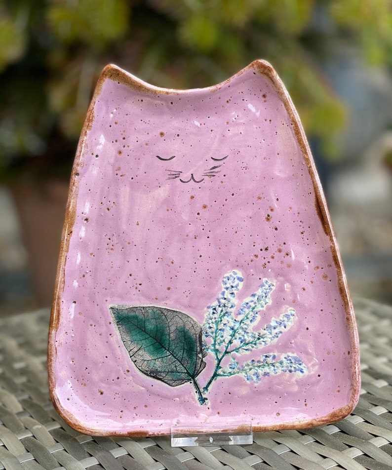 May include: A pink ceramic cat-shaped dish with a leafy design and a small flower sprig. The dish has a speckled glaze and a brown rim.