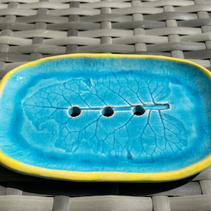 Könnte beinhalten: Eine blaue Keramik-Seifenschale mit gelbem Rand und einem Blattdesign. Die Schale hat vier Abflusslöcher.