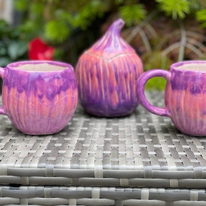 Peut inclure: Deux tasses en céramique violettes et roses avec un sucrier en céramique en forme de figue assorti. Les tasses et le sucrier ont un motif tourbillonné en tie-dye.