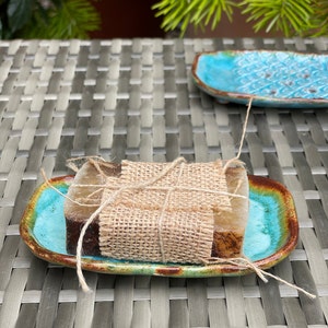 May include: Two bars of soap wrapped in burlap twine and placed on a turquoise and brown ceramic soap dish.