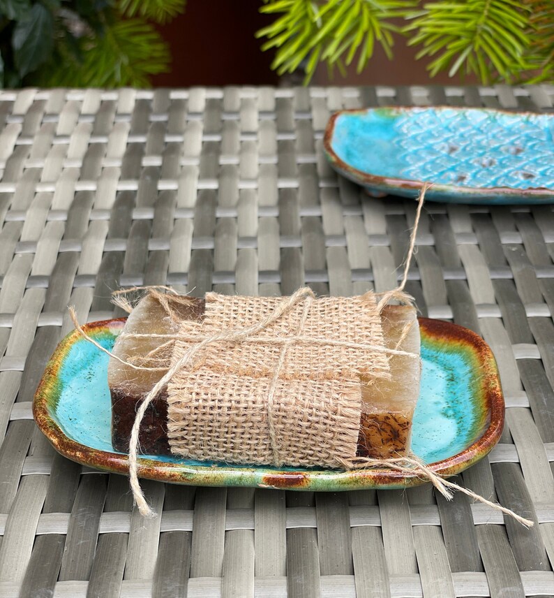 May include: Two bars of soap wrapped in burlap twine and placed on a turquoise and brown ceramic soap dish.