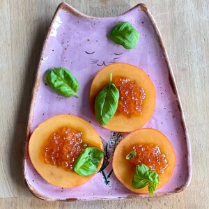 May include: Three slices of peach topped with red caviar and fresh basil leaves on a pink cat-shaped plate.