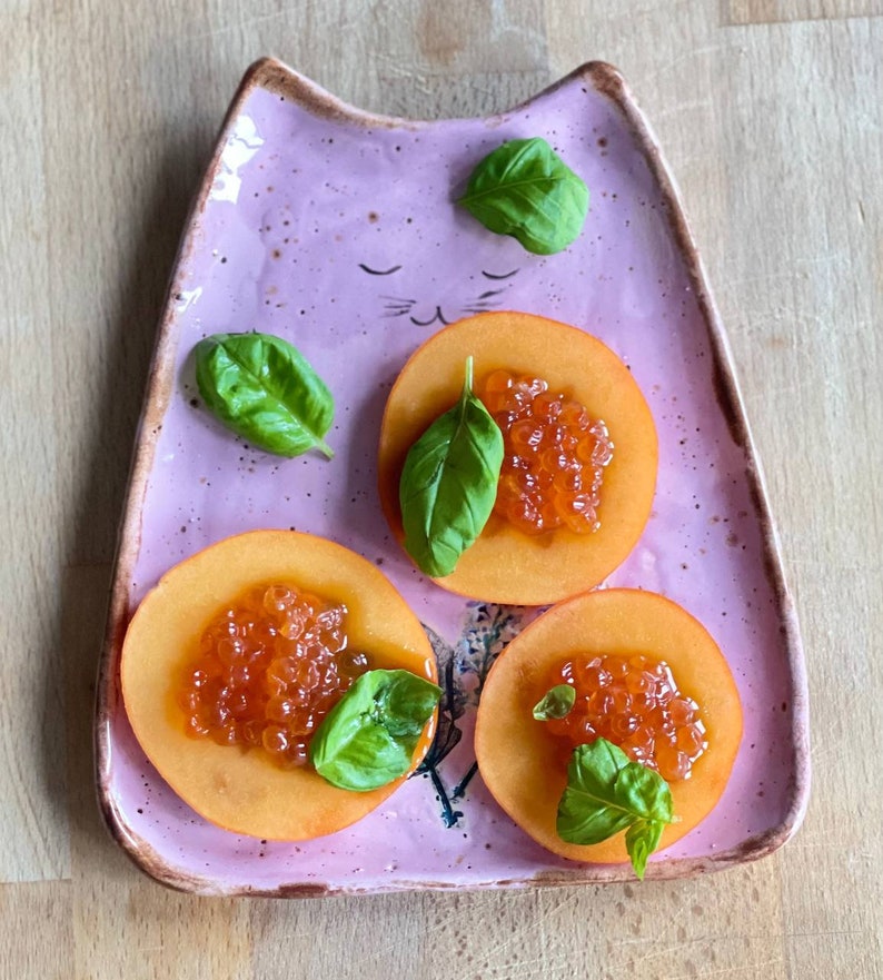 May include: Three slices of peach topped with red caviar and fresh basil leaves on a pink cat-shaped plate.