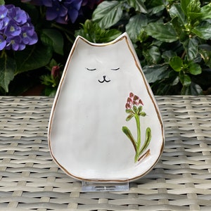 Peut inclure: Plat en céramique blanche en forme de chat avec un bord doré et une fleur rose peinte sur le côté. Le chat a les yeux fermés et un petit nez.