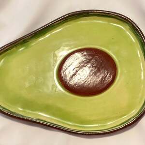 May include: A green ceramic avocado-shaped serving dish with a brown pit in the center. The dish is glazed and has a smooth finish.