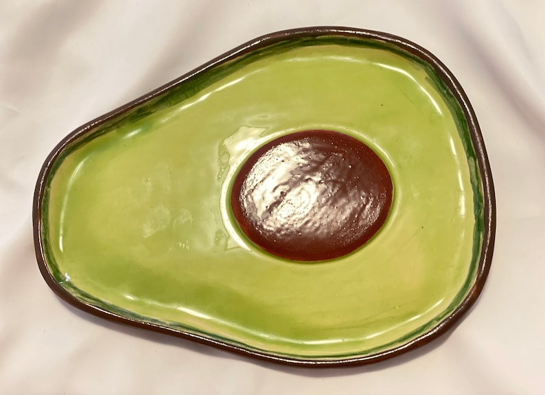 May include: A green ceramic avocado-shaped serving dish with a brown pit in the center. The dish is glazed and has a smooth finish.