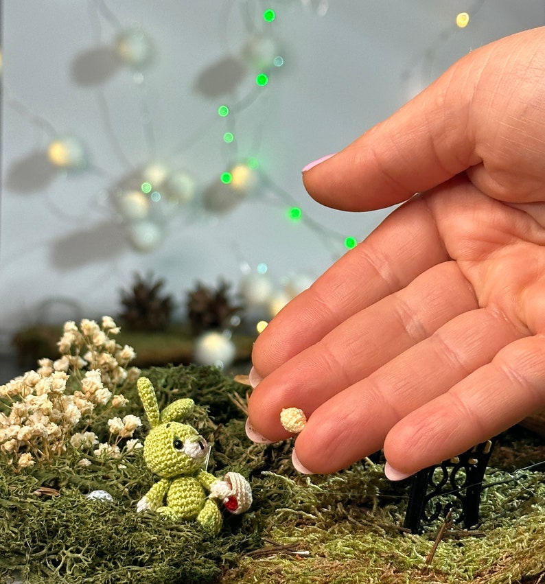 May include: A miniature crocheted green bunny holding a basket with a red item, set in a mossy scene with white flowers and a tiny black bench. A hand holds a small, round, beige item. Fairy garden theme.