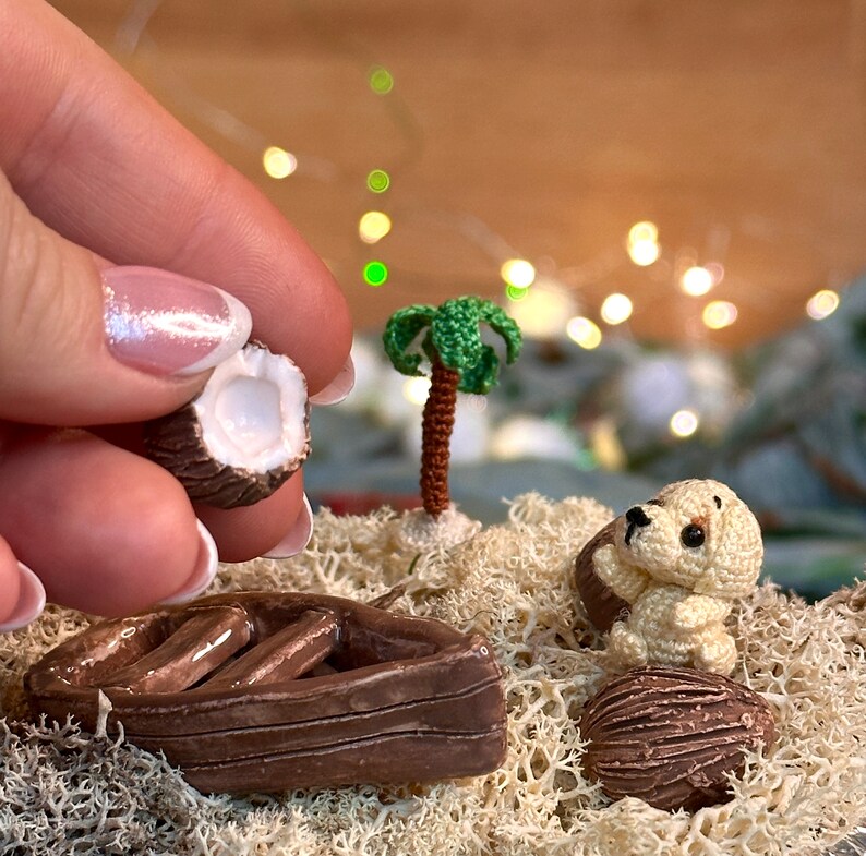 May include: A miniature crochet scene featuring a half coconut held by a hand, a small palm tree, a boat, a puppy, and a whole coconut. The scene is set on a bed of light green moss, with a blurred background of lights.