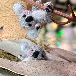 May include: Two handmade crochet koala bears, one hanging from a branch and the other resting on a leaf. The koalas are gray with black spots, pink cheeks, and black eyes. The background includes dried leaves and a blurred bokeh effect.