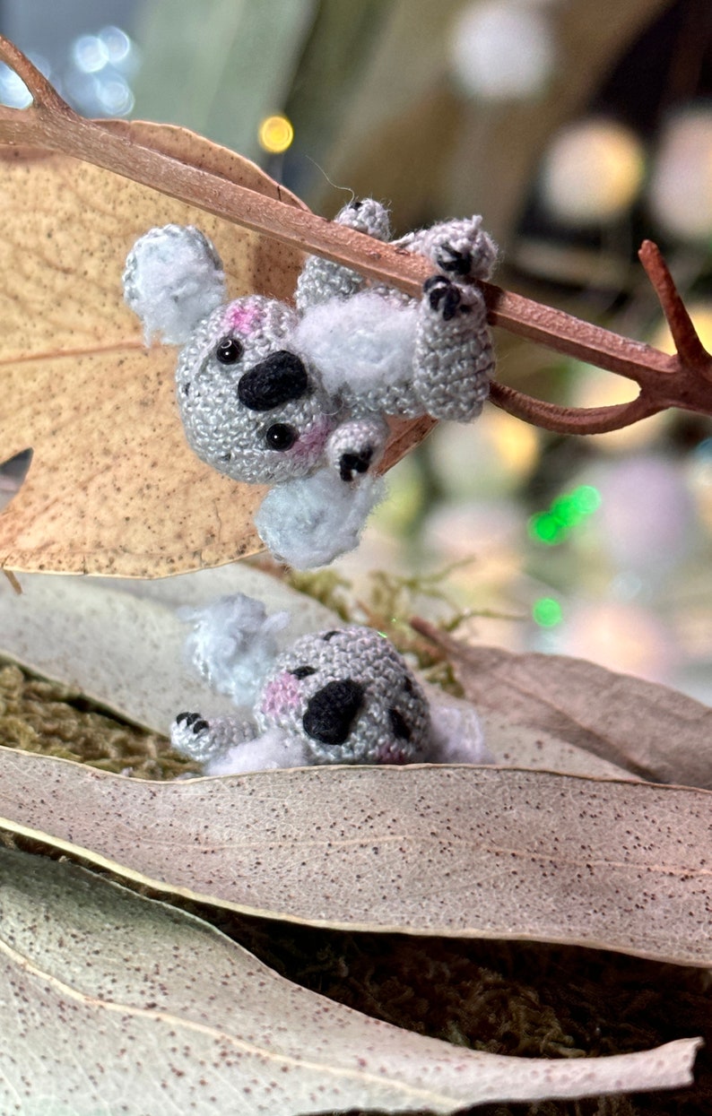 May include: Two handmade crochet koala bears, one hanging from a branch and the other resting on a leaf. The koalas are gray with black spots, pink cheeks, and black eyes. The background includes dried leaves and a blurred bokeh effect.