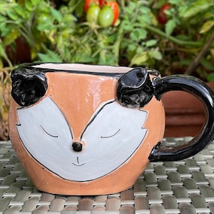 Peut inclure: Une tasse en céramique avec un motif de visage de renard. La tasse est de couleur pêche avec des oreilles noires et un visage blanc. La tasse a une poignée noire.