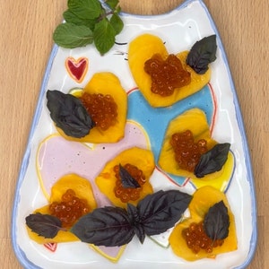 May include: A white ceramic plate shaped like a cat with a blue rim, decorated with pink and yellow hearts. The plate is filled with heart-shaped slices of yellow fruit topped with red caviar and garnished with green and purple basil leaves.