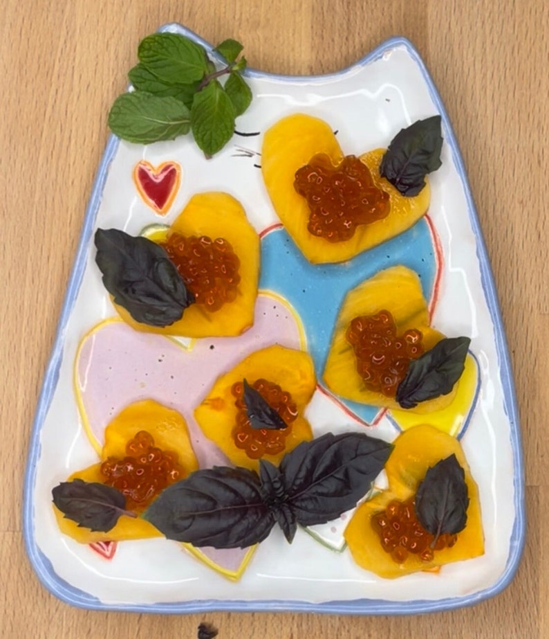May include: A white ceramic plate shaped like a cat with a blue rim, decorated with pink and yellow hearts. The plate is filled with heart-shaped slices of yellow fruit topped with red caviar and garnished with green and purple basil leaves.