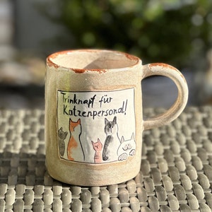 Könnte beinhalten: Eine beige Keramiktasse mit braunem Rand und Henkel. Die Tasse hat eine schwarz-weiße Illustration von fünf Katzen und den Text "Trinknapf für Katzenpersonal!"
