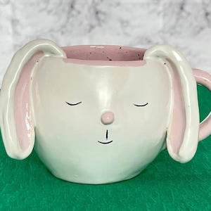 Può includere: Tazza in ceramica bianca a forma di coniglio con orecchie rosa e bordo rosa. Il coniglio ha gli occhi chiusi e un piccolo naso rosa.