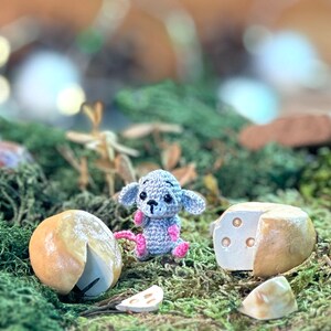 May include: A crocheted gray mouse with pink accents sits near miniature cheese wheels and slices on a bed of green moss. The scene evokes a whimsical, handcrafted aesthetic, perfect for a nursery or a unique gift.