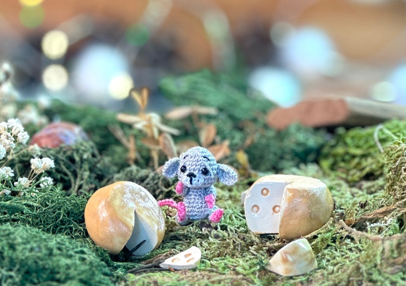 May include: A crocheted gray mouse with pink accents sits near miniature cheese wheels and slices on a bed of green moss. The scene evokes a whimsical, handcrafted aesthetic, perfect for a nursery or a unique gift.
